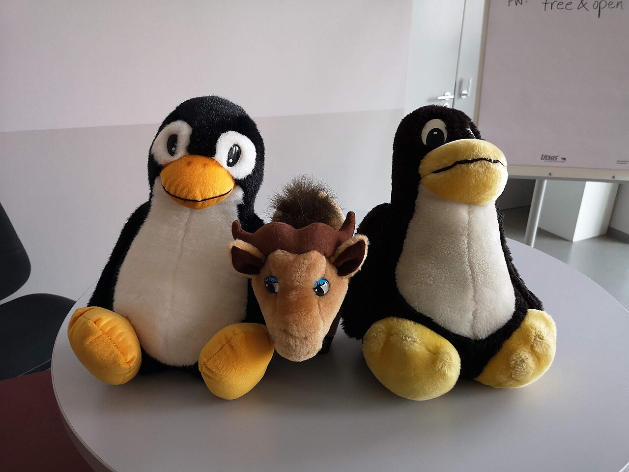 Deux peluches Tux, mascotte de Linux, entourant une peluche GNU sur une table, en 2019.